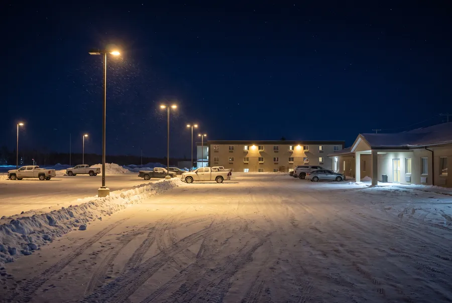 Generated scene: a snow-covered parking lot at night, lamp posts throwing pools of warm light, pickup trucks, a two-storey hotel with lit windows and a low building with a covered entrance.