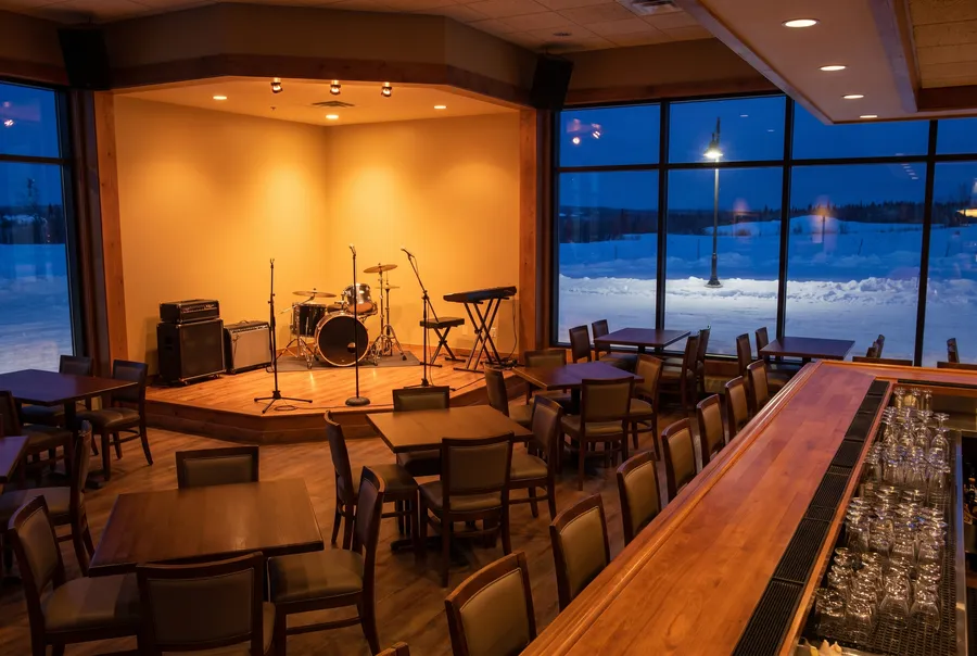 Generated scene: an empty lounge with a small stage, a drum kit, amplifiers and a keyboard under warm lights, wooden tables, a long bar and a snowy night through tall windows.